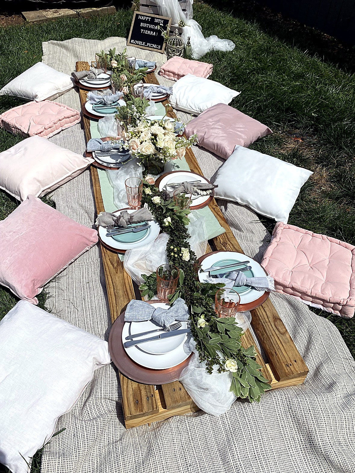 LoveLe Picnics 30th birthday setup — long low table with blush pillows, floral garland and place settings on the lawn